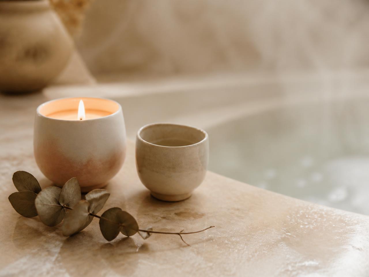 A lit ceramic candle and dried eucalyptus on a warm stone bath ledge